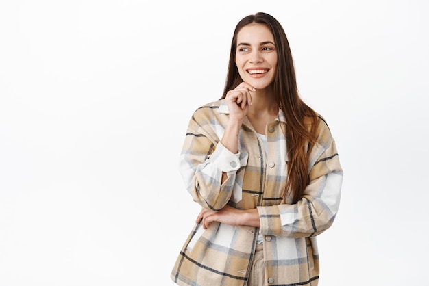 Free photo image of stylish young professional woman touching face smiling and laughing selfassured looking aside at copy space promotional text on the left standing over white background