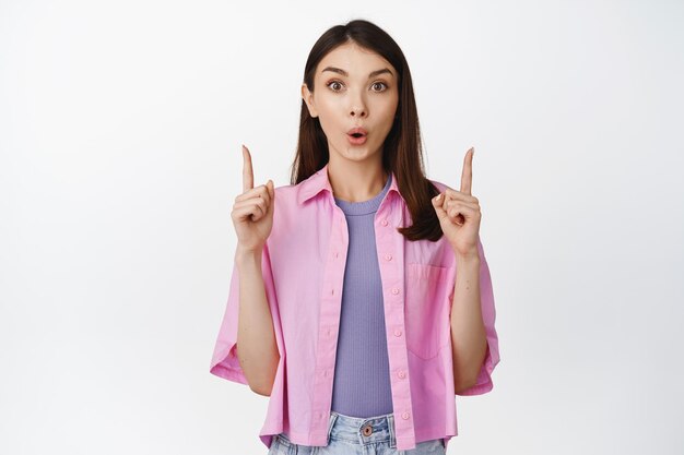 Image of surprised brunette girl with curious face expression pointing fingers up interested about sale discounts standing over white background