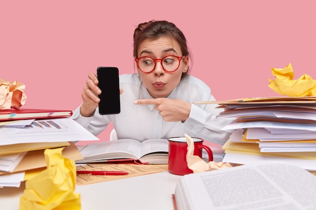 Free photo image of surprised european woman has shocked expression, has folded lips, shows something on blank space, carries modern cell phone