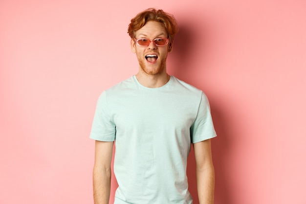 Free Photo image of surprised redhead man on vacation, wearing sunglasses with summer t-shirt, open mouth and saying wow amazed, standing over pink background.