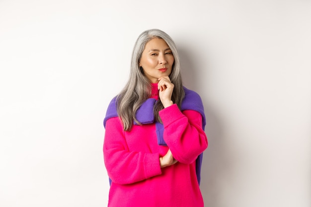 Image of thoughtful asian senior woman squinting suspicious at camera, having assumption, standing over white background and thinking