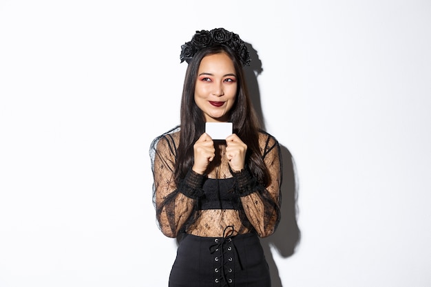 Free photo image of thoughtful asian woman in black gothic dress have a plan, holding credit card, smiling pleased at upper left corner.