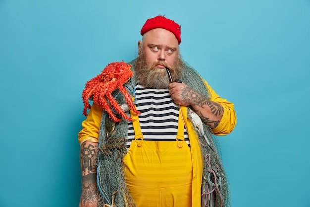 Free Photo image of thoughtful man has marine occupation, smokes pipe with pensive sad expression, poses with fishing equipment, carries octopus, thinks about next sea voyage or adventure