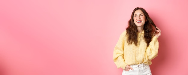 Free photo image of young 20s woman laughing and playing with her hair looking aside at upper left corner promotion standing happy against pink background