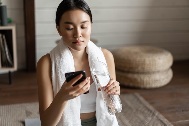 Free photo image of young asian fitness girl with fit body towel wrapped around neck looking at mobile phone ap...