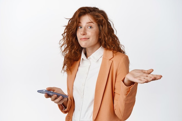 Image of young business woman in suit holding mobile phone and shrugging clueless standing puzzled and smiling to apologize white background