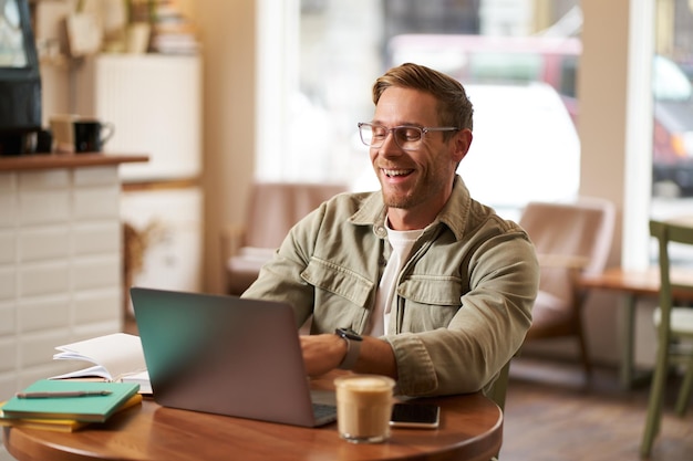 Image of young digital nomad man in glasses sits in cafe works from coffee shop uses laptop in