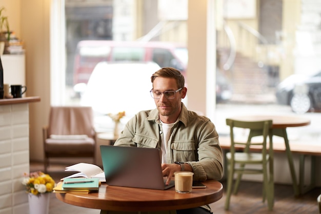 Free photo image of young digital nomad man in glasses sits in cafe works from coffee shop uses laptop in