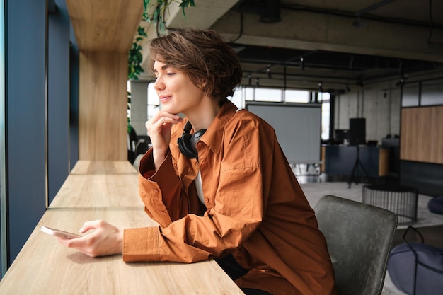 Image of young digital nomad student at coworking space sitting at table with mobile phone looking