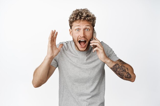 Image of young man talks on phone receives a phone call and looking with disbelief shouting at camera with excited face expression white background