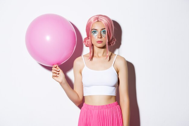 Image of young stylish girl holding pink balloon, wearing party wig and celebrating holiday, standing.