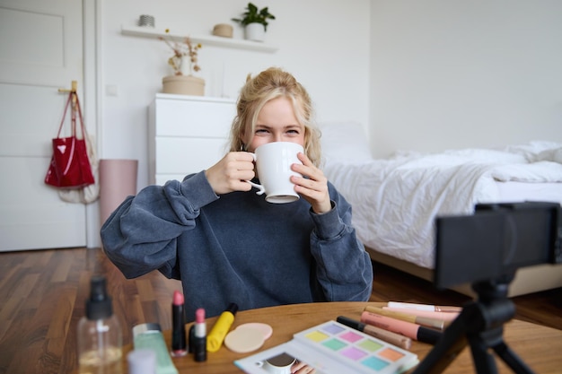 Free photo image of young woman makeup vlogger sitting in bedroom with digital camera drinking tea and talking