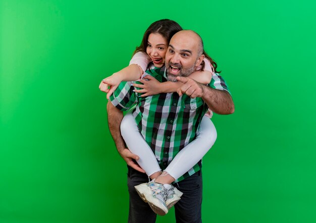 Impressed adult couple man holding woman on his back both looking and pointing at side isolated on green wall with copy space