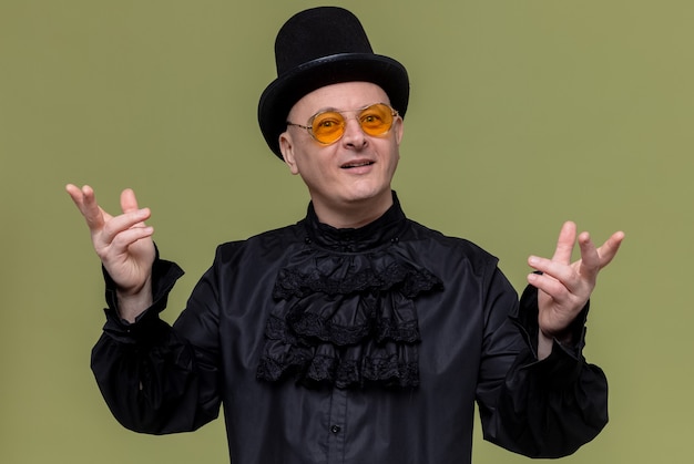 Free photo impressed adult man with top hat and with sunglasses in black gothic shirt keeping hands open and looking