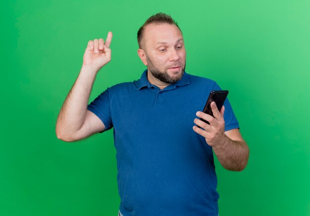 Impressed adult slavic man holding and looking at mobile phone raising finger isolated on green wall