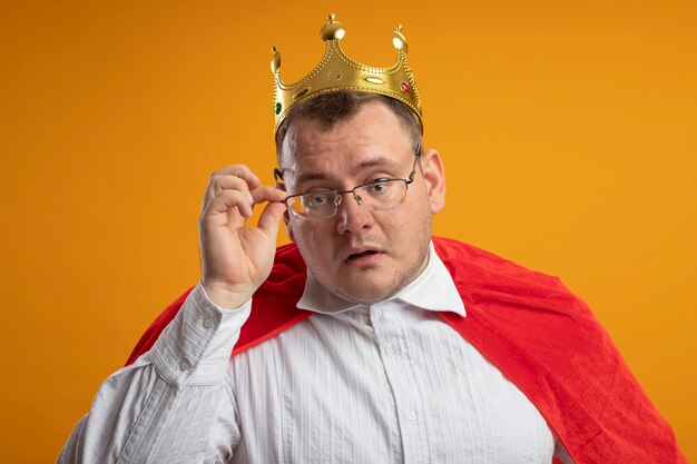 Impressed adult slavic superhero man in red cape wearing glasses and crown grabbing glasses  isolated on orange wall