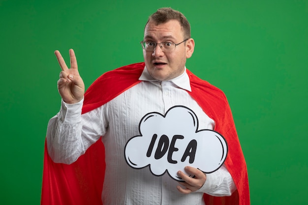 Free photo impressed adult slavic superhero man in red cape wearing glasses holding idea bubble holding idea bubble doing peace sign isolated on green wall