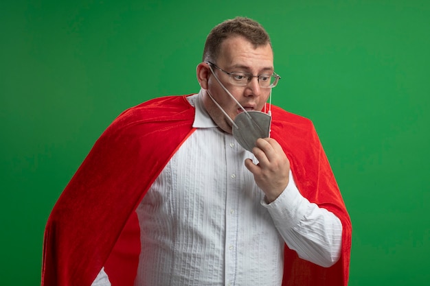 Free photo impressed adult superhero man in red cape wearing glasses and protective mask looking down trying to take off his mask isolated on green wall