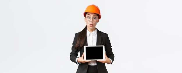 Impressed asian female construction engineer businesswoman in safety helmet and suit showing chart introduce digram at digial tablet screen standing amazed over white background