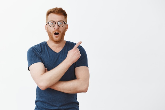 Free photo impressed attractive caucasian guy with bristle in glasses and blue t-shirt, pointing at upper right corner, gasping and dropping haw from astonishment