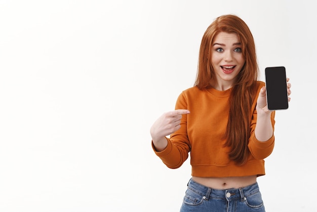 Free photo impressed attractive millennial redhead female in cropped sweater holding smartphone pointing mobile screen and smiling amused as talking about new application tracker or online shopping