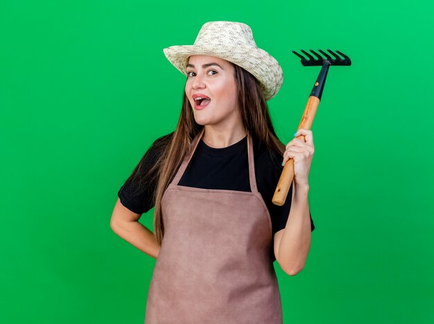 Impressed beautiful gardener girl in uniform wearing gardening hat raising rake isolated on green