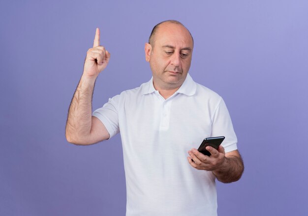 Impressed casual mature businessman holding and looking at mobile phone raising finger isolated on purple background