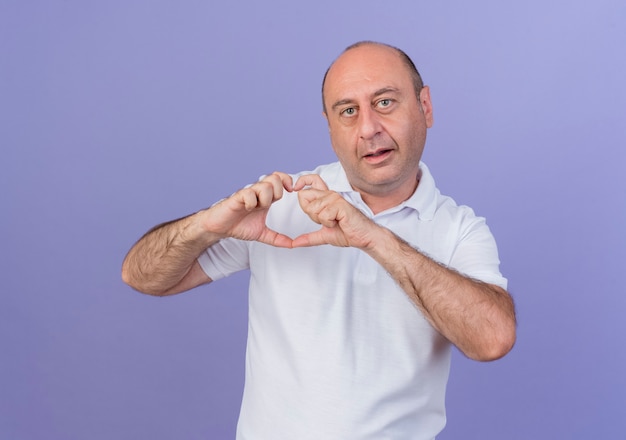 Impressed casual mature businessman looking at camera and doing heart sign isolated on purple background