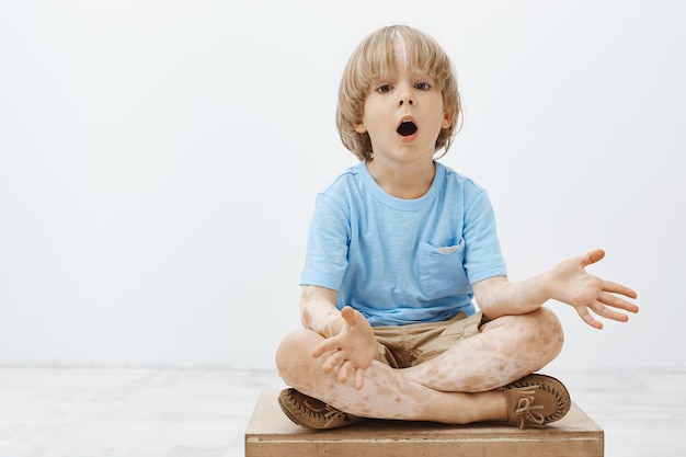 impressed curious blond child with vitiligo sitting with crossed feet and dropping jaw, spreading palms