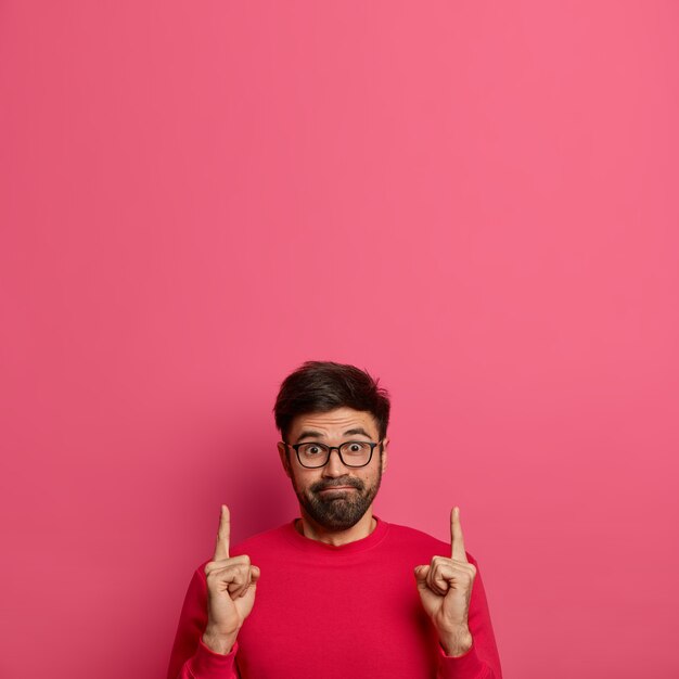 Impressed curious man presses lips, advertises product, points above with both index fingers