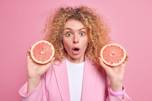 Free photo impressed european woman holds two halves of fresh grapefruit surprised to find out about useful substances which this citrus fruit contains has curly hair isolated over pink wall. dieting