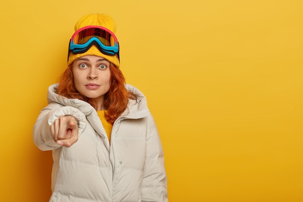 Free photo impressed ginger woman snowboarder points into camera, dressed in outerwear, protective snowboarding goggles, isolated over yellow background. winter resort concept