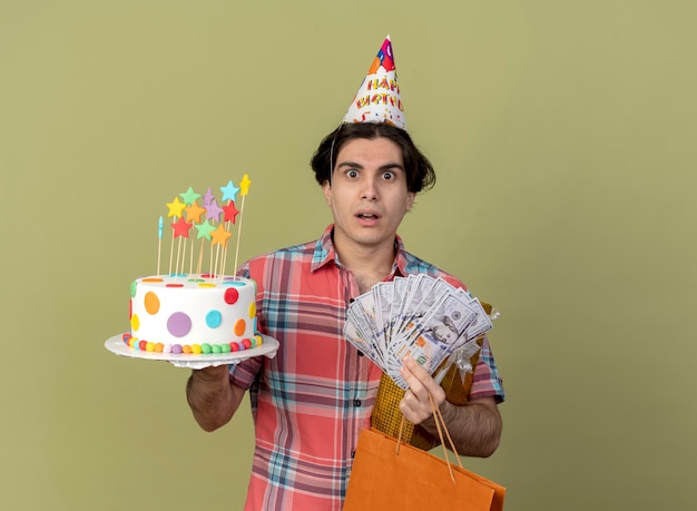 Impressed handsome caucasian man wearing birthday cap holds gift box paper shopping bag money and birthday cake 