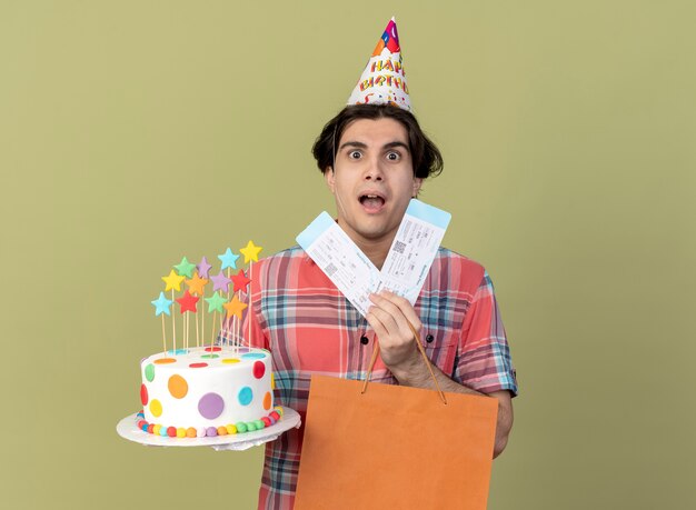 Impressed handsome caucasian man wearing birthday cap holds paper shopping bag air tickets and birthday cake 