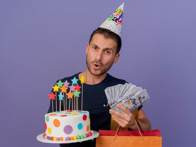 Impressed handsome man wearing birthday cap holds birthday cake paper shopping bag gift box and money isolated on purple wall