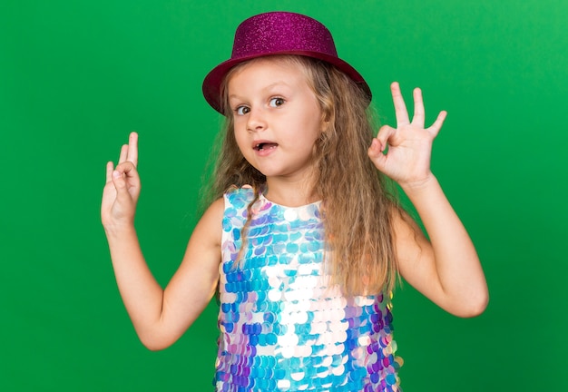 impressed little blonde girl with purple party hat gesturing ok sign isolated on green wall with copy space