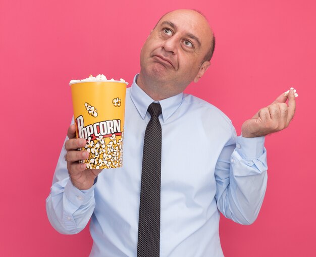 Impressed looking at side middle-aged man wearing white t-shirt with tie holding bucket of popcorn with popcorn piece isolated on pink wall