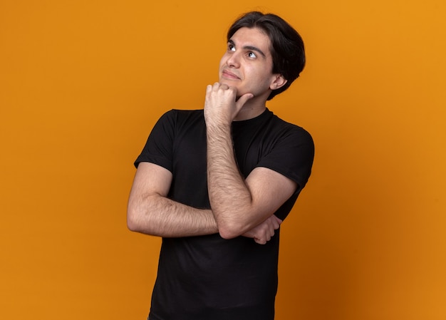 Impressed looking at side young handsome guy wearing black t-shirt putting fist under chin isolated on orange wall