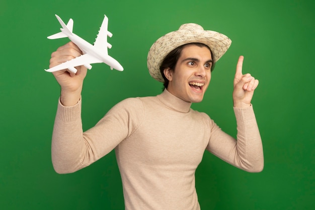 Free Photo impressed looking at side young handsome guy wearing hat and raising toy airplane points at up isolated on green wall