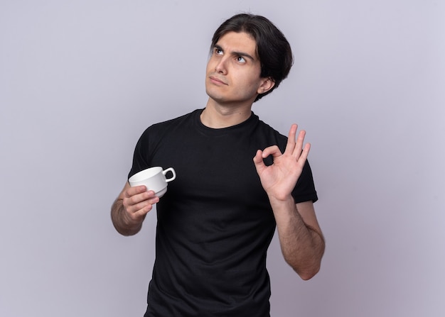 Impressed looking up young handsome guy wearing black t-shirt holding cup of coffee showing okay gesture isolated on white wall