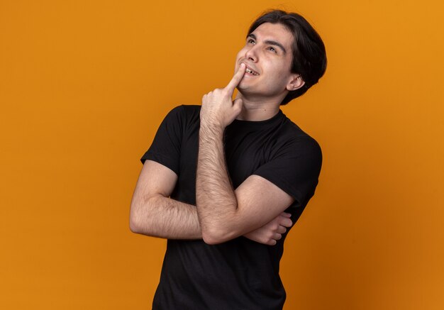 Impressed looking up young handsome guy wearing black t-shirt putting finger on mouth isolated on orange wall