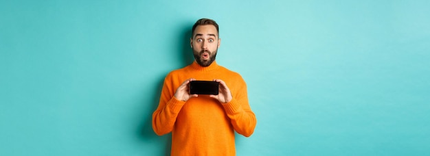 Impressed man showing smartphone screen stare at camera amazed demonstrate display standing over lig