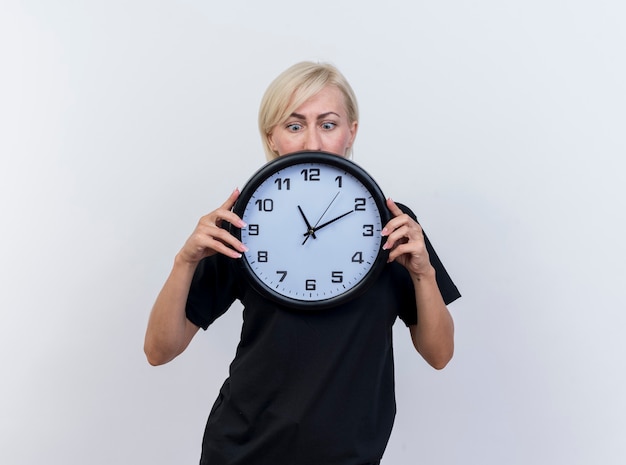 Impressed middle-aged blonde slavic woman holding and looking at clock isolated on white background with copy space