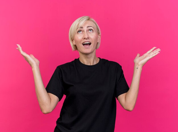 Impressed middle-aged blonde slavic woman looking up showing empty hands isolated on pink wall