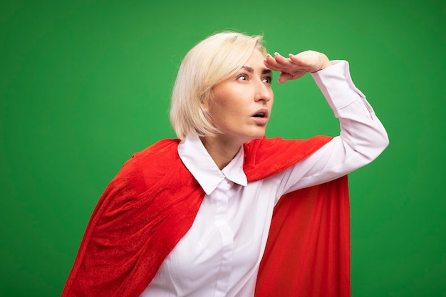 Free photo impressed middle-aged blonde superhero woman in red cape keeping hand on forehead looking at side into distance