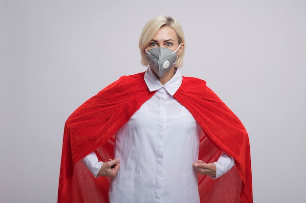 Impressed middle-aged blonde superhero woman in red cape wearing protective mask standing like superman  isolated on white wall