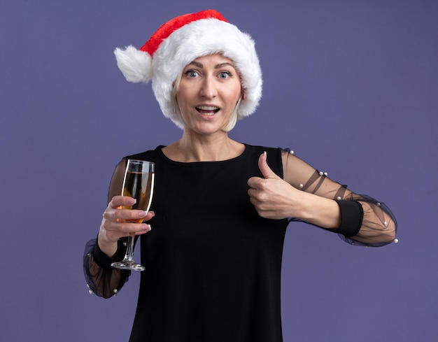 Free photo impressed middle-aged blonde woman wearing christmas hat looking at camera holding glass of champagne showing thumb up isolated on purple background