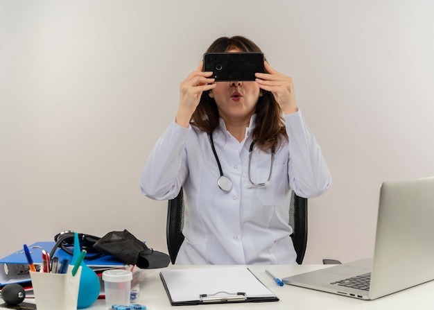 Free photo impressed middle-aged female doctor wearing medical robe and stethoscope sitting at desk with medical tools clipboard and laptop holding and hiding behind mobile phone isolated