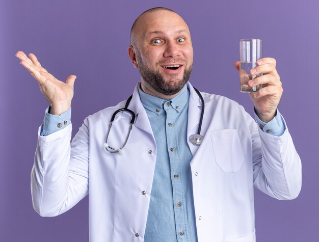 Impressed middle-aged male doctor wearing medical robe and stethoscope holding glass of water  showing empty hand isolated on purple wall