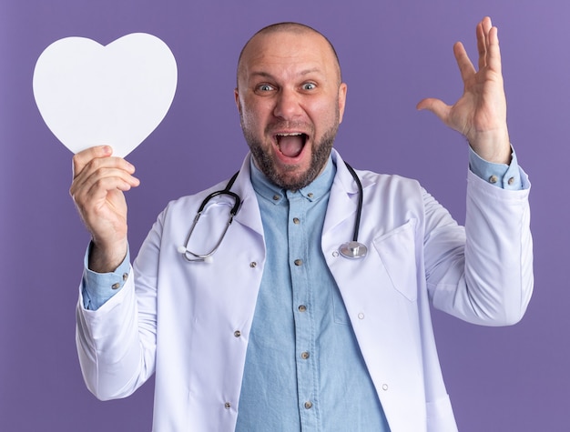 Free photo impressed middle-aged male doctor wearing medical robe and stethoscope holding heart shape raising hand